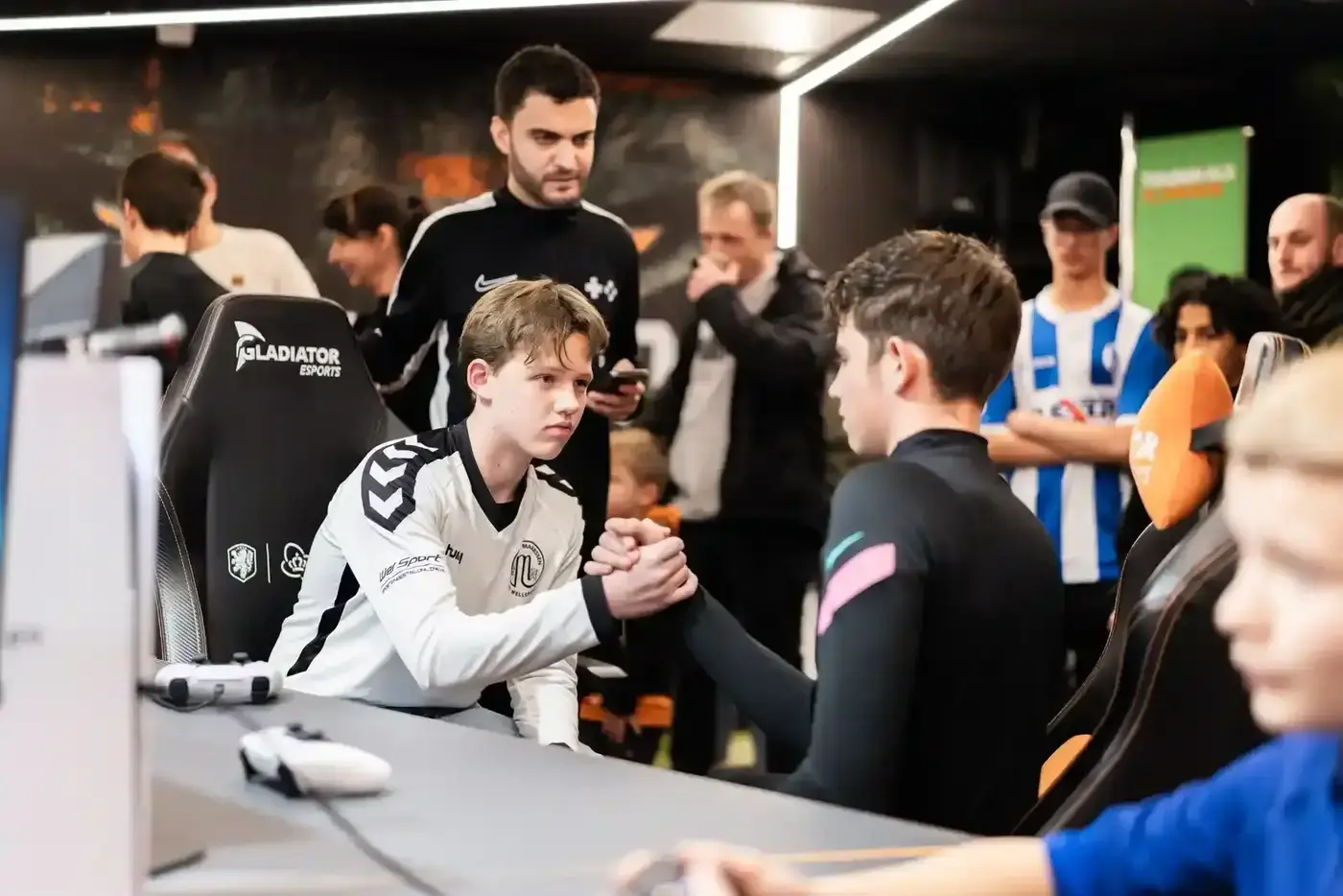 Twee jonge spelers schudden handen na een FIFA-wedstrijd terwijl toeschouwers en begeleiders toekijken
