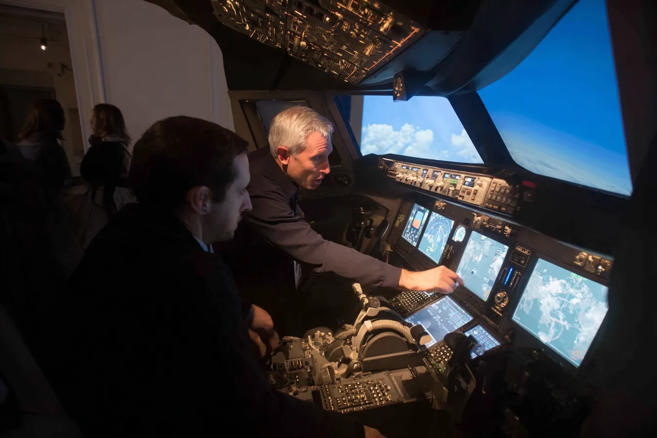 Instructeur staat naast deelnemer in vluchtsimulator en geeft aanwijzingen tijdens een landing
